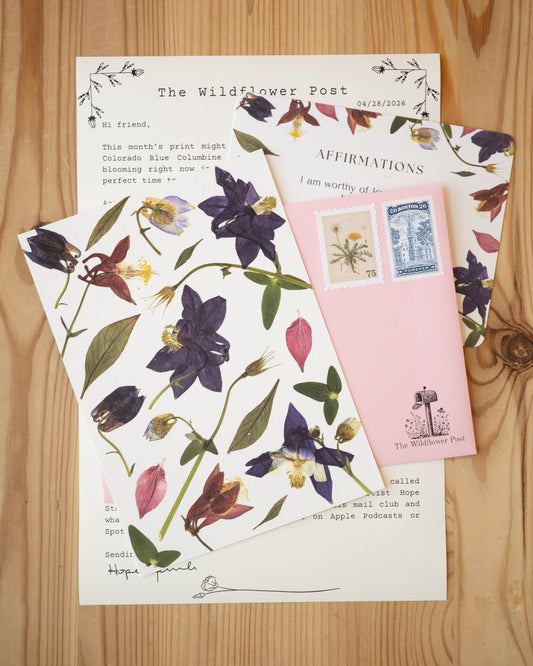 Art print of pressed columbine with letter and affirmation card on table.