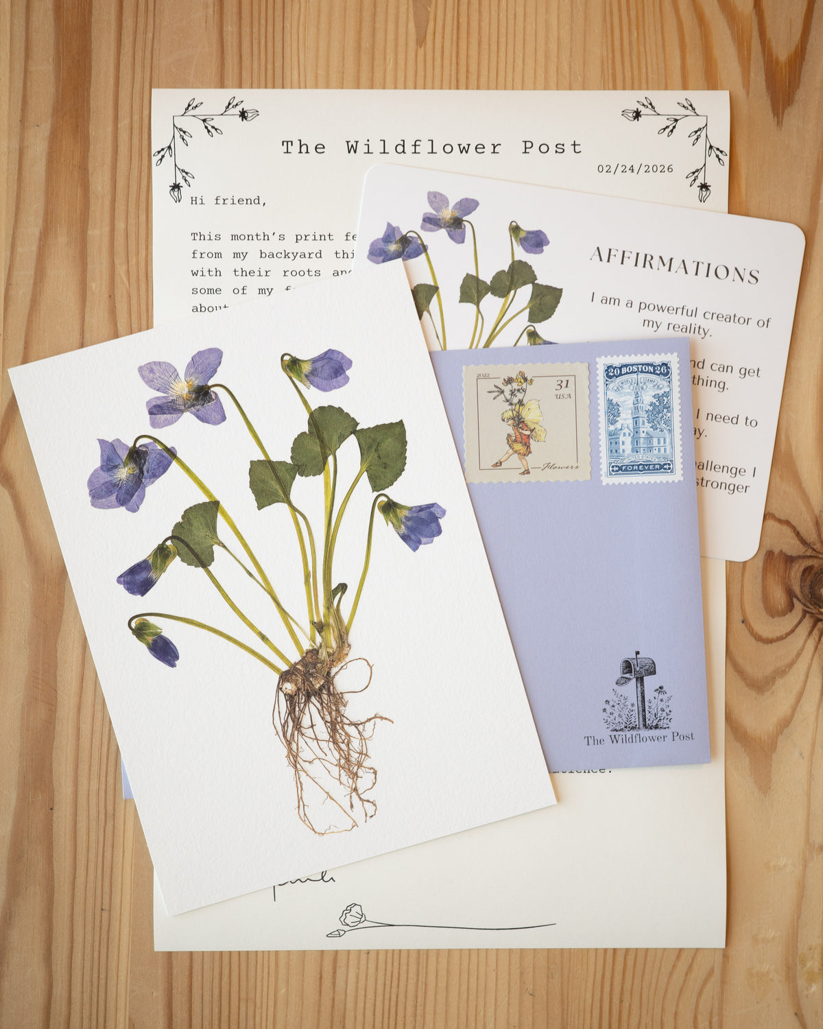 Art print of pressed violet with letter and affirmation card on table. 