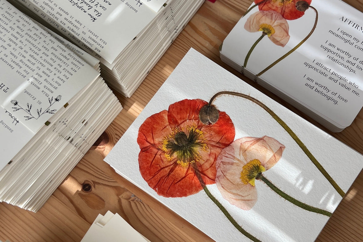 A styled flat-lay of fine art print featuring pressed poppy flower, arranged on a wooden table alongside stacks of affirmation cards and red envelopes.