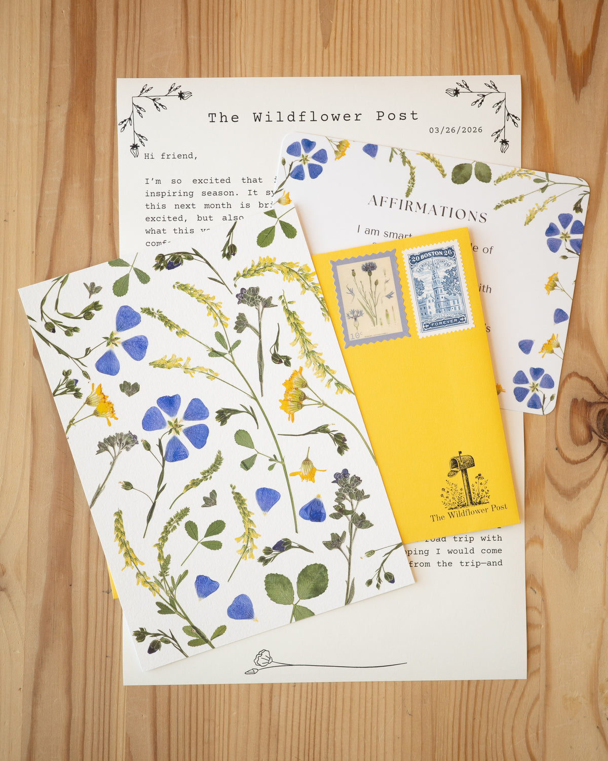Art print of pressed blue and yellow wildflowers with letter and affirmation card on table.