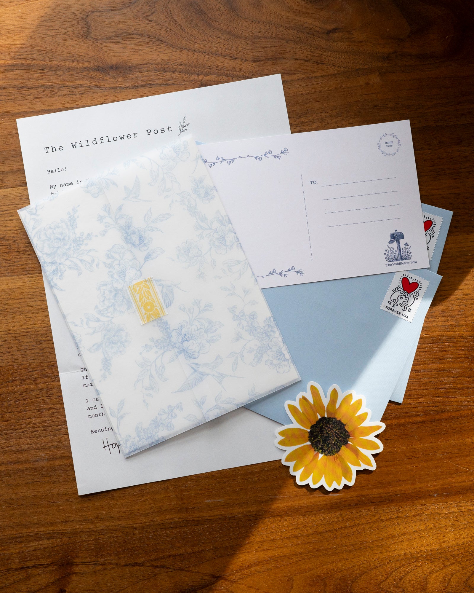 Contents of snail mail club on desk including letter, postcard, envelope, sticker, and art pritnt in vellum jacket.