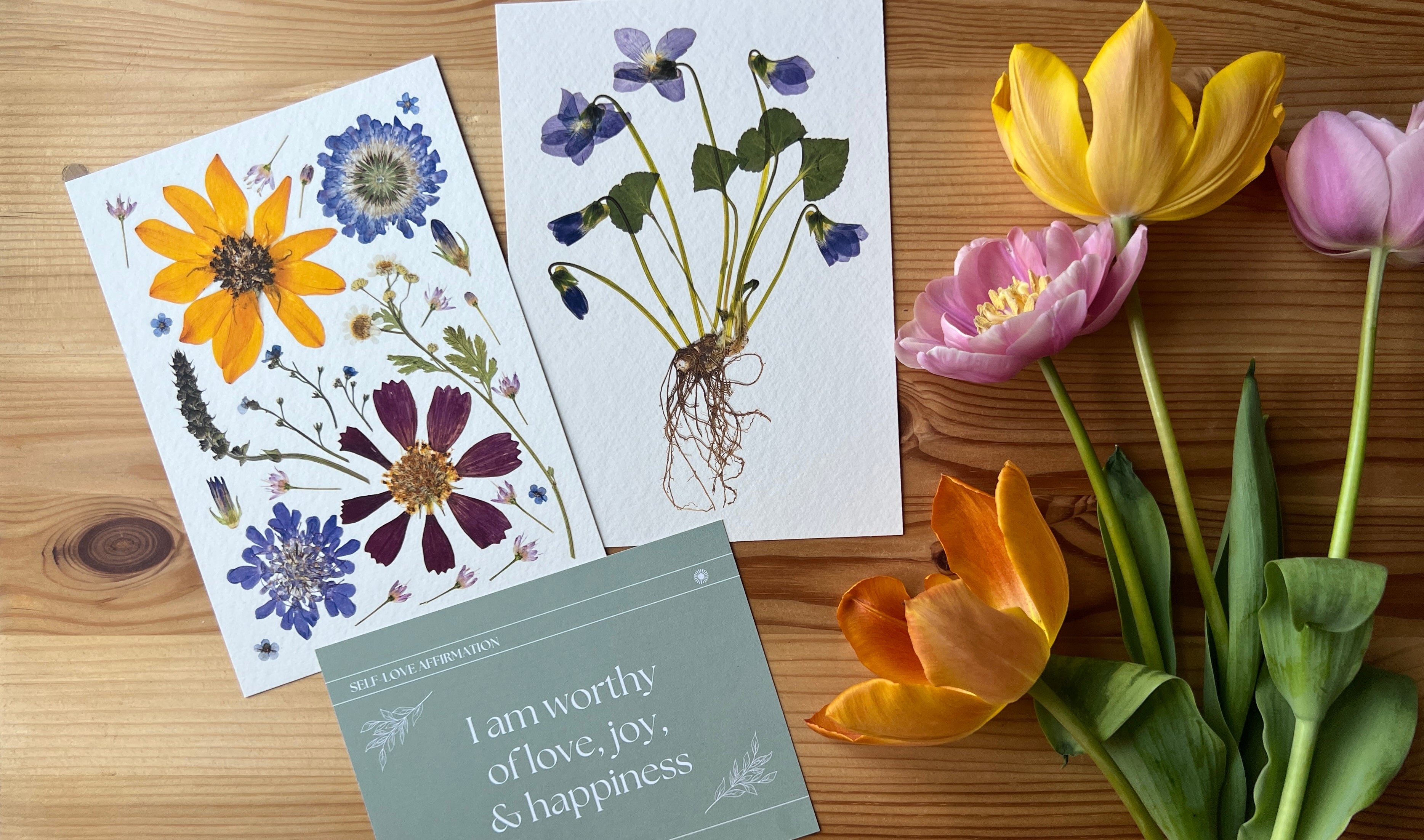 Pressed flower art prints laying on table with fresh yellow and pink tulips and a green card with an affirmation on it.