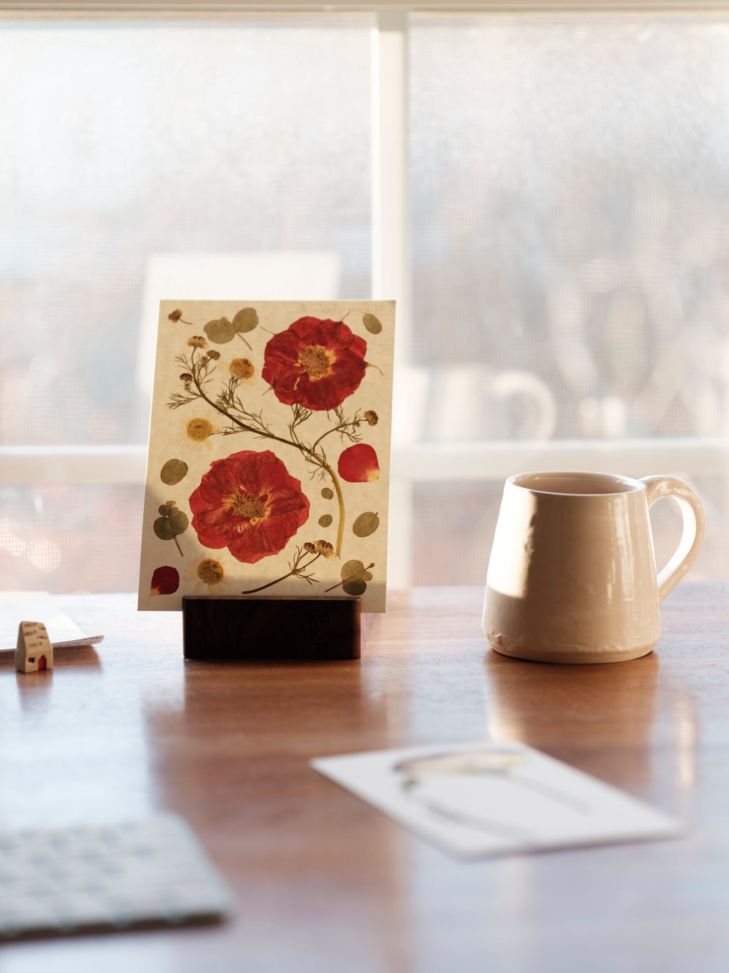 Small pressed flower art print from monthly print club displayed on desk with coffee mug in early morning light.