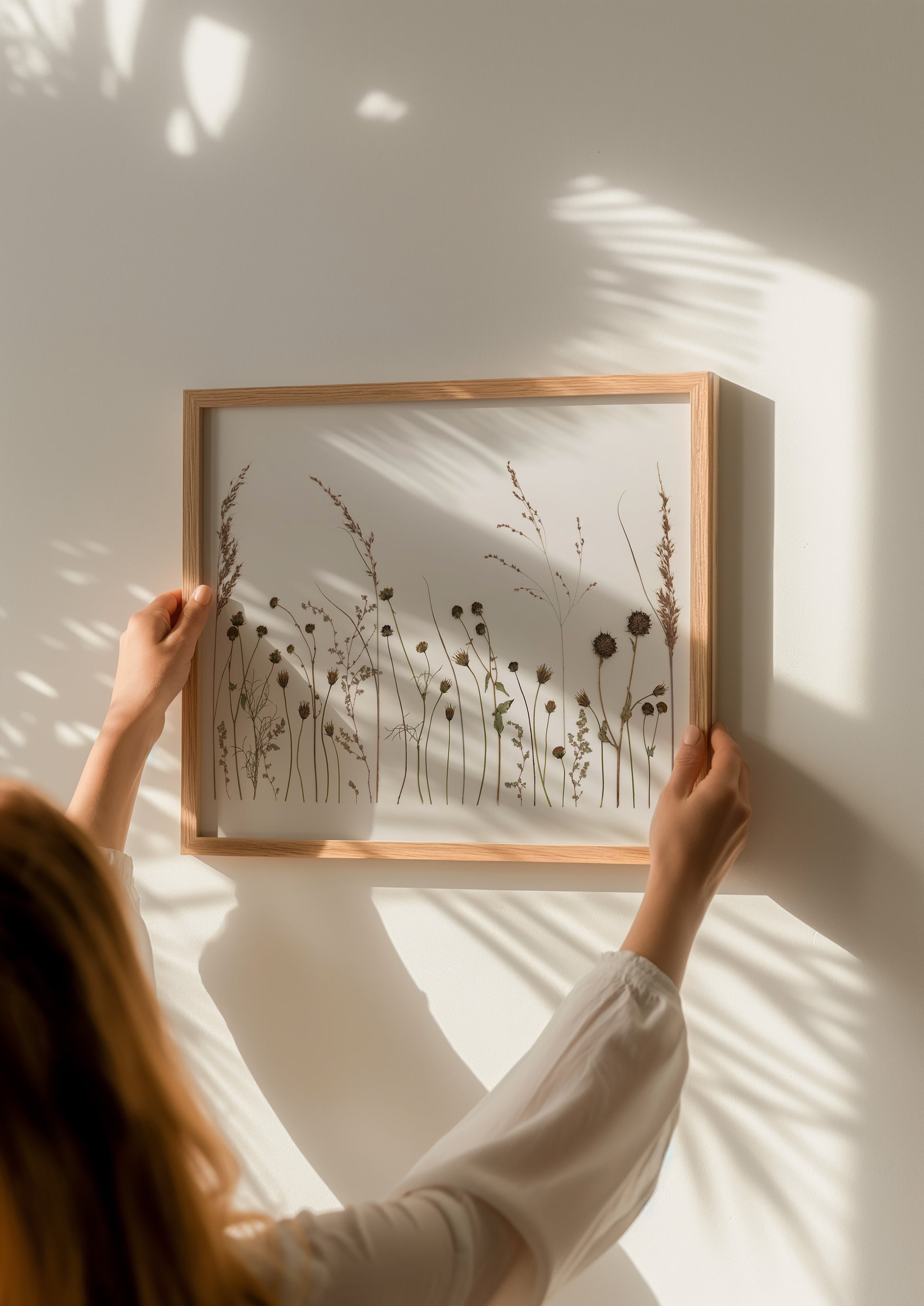 Person holding a framed art print of pressed tall grass flowers against a light wall.